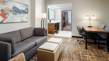 Quarto de hotel moderno com sofá cinzento, um banquinho bege, tapete estampado, mesa de jantar com cadeiras, luminária e um quadro abstrato grande na parede.