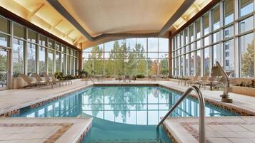 An indoor swimming pool with clear glass walls, lounge chairs, and a tiled deck.