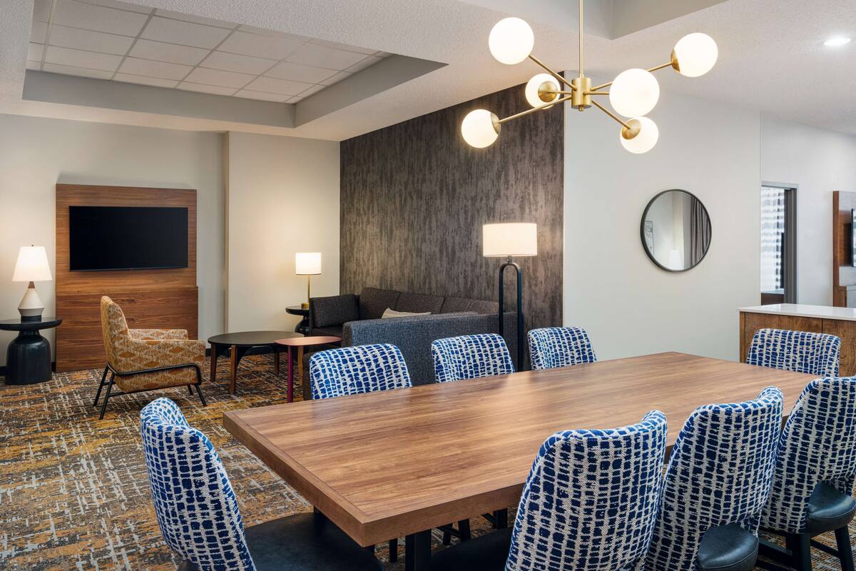 Une salle de conférence d’hôtel avec une grande table en bois, des chaises à motifs, un canapé, un téléviseur et un lustre moderne.