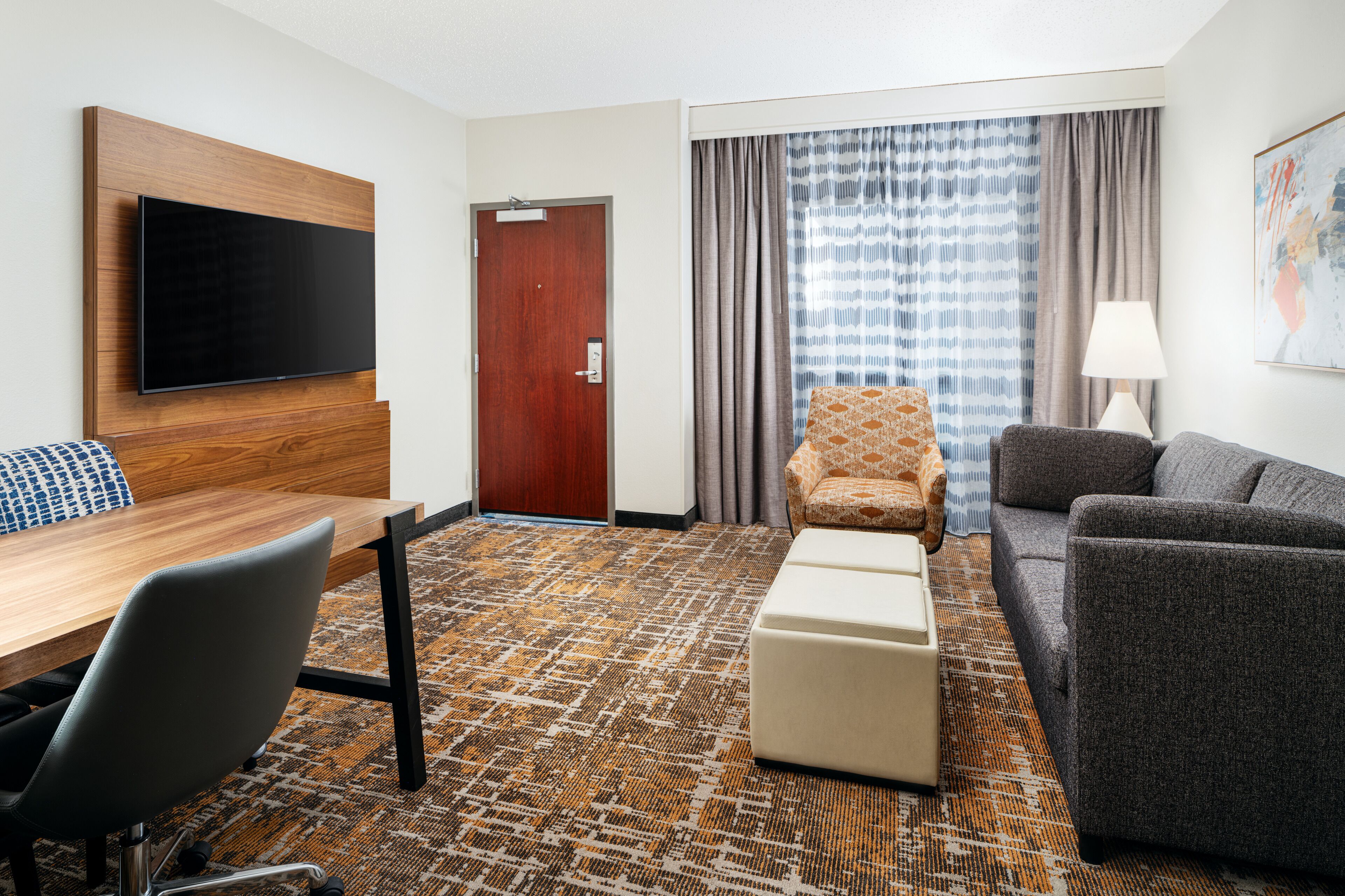 Habitación de hotel con televisión de pantalla plana montada en un panel de madera, un escritorio de madera con una silla, un sofá, una alfombra estampada y una puerta que ofrece vistas del exterior a través de cortinas.