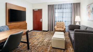 A hotel room with a flat-screen TV mounted on a wooden panel, a wooden desk with a chair, a sofa, a patterned carpet, and a door with a view of the outside through curtains.