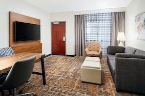 Hotelkamer met flatscreen televisie, bevestigd aan een houten paneel, een houten bureau met stoel, een bank, een kamerbreed tapijt en een deur met uitzicht buiten door gordijnen.