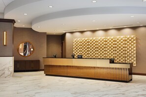 Un espace d’accueil d’hôtel au design moderne, avec un sol en marbre, un comptoir d’accueil en bois, un miroir et un mur à texture.
