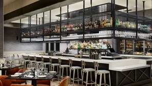 Een moderne barruimte met meerdere flessenrekken en glazen, een toonbank met barbenodigdheden, en een zithoek met barstoelen en tafels.