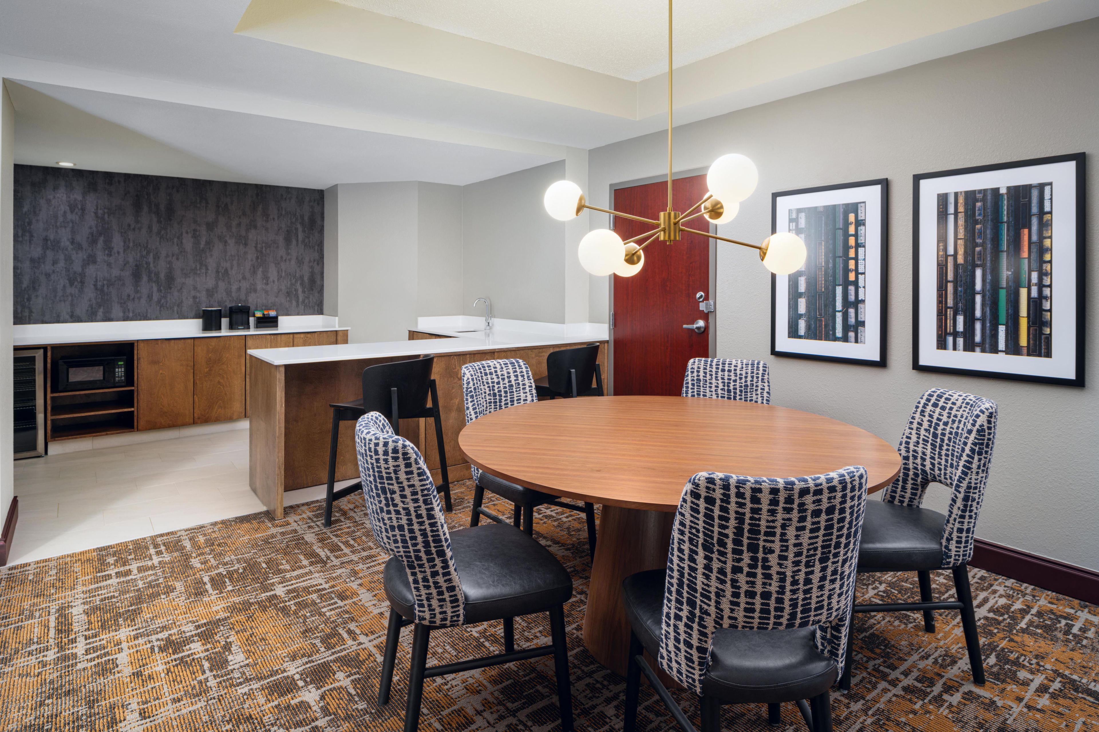 A modern hotel room with a round dining table, black upholstered chairs, a kitchen area, and a large window.