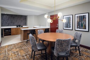 Una habitación de hotel moderna con una mesa de comedor redonda, sillas tapizadas en negro, una zona de cocina y un ventanal grande.