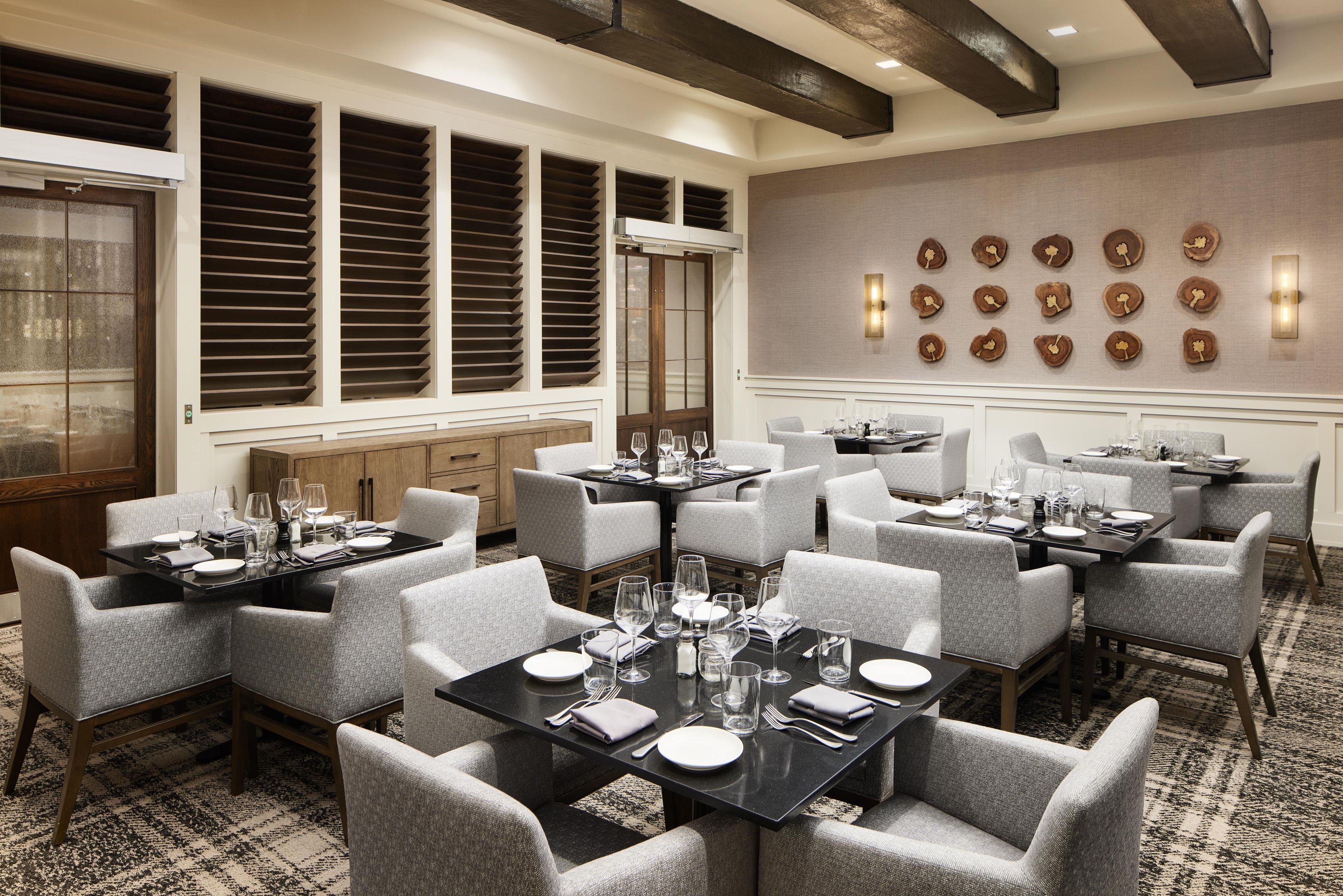 A hotel dining area with multiple tables set for dining, a large window with blinds, and a wall with decorative wood inlays.
