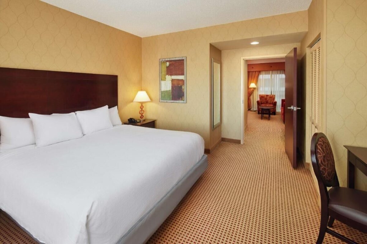 Ein Hotelzimmer mit einem großen Bett, einem Schreibtisch, einem Stuhl, einer Lampe und einer Tür zu einem weiteren Raum.
