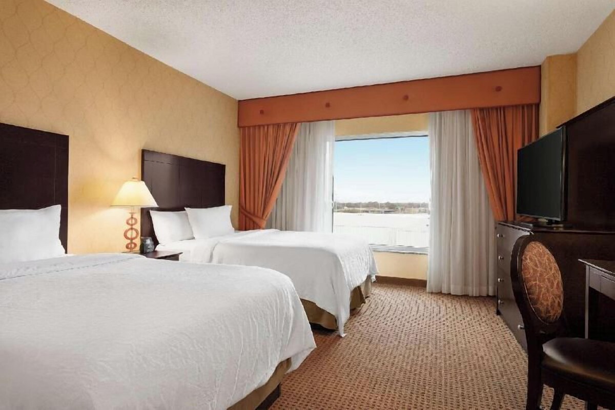 A hotel room with two beds, a television, a desk, and a view of the outdoors.