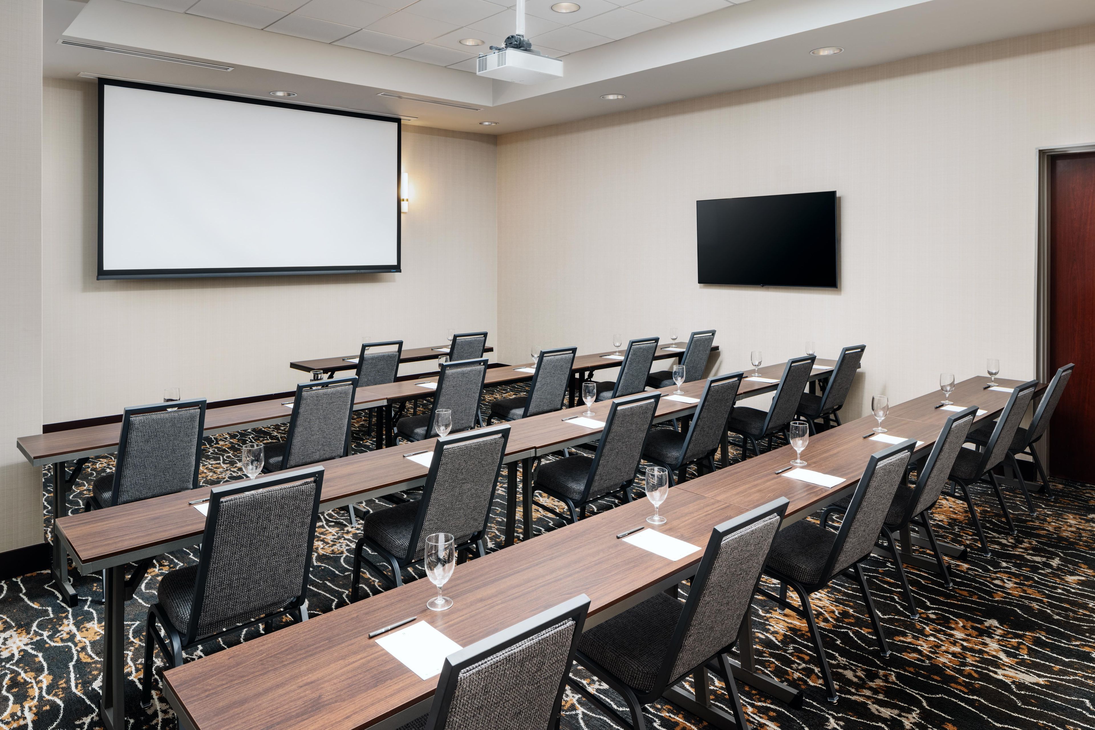 Un salón de conferencias con una pantalla de proyector, un televisor de pantalla plana y piso alfombrado.