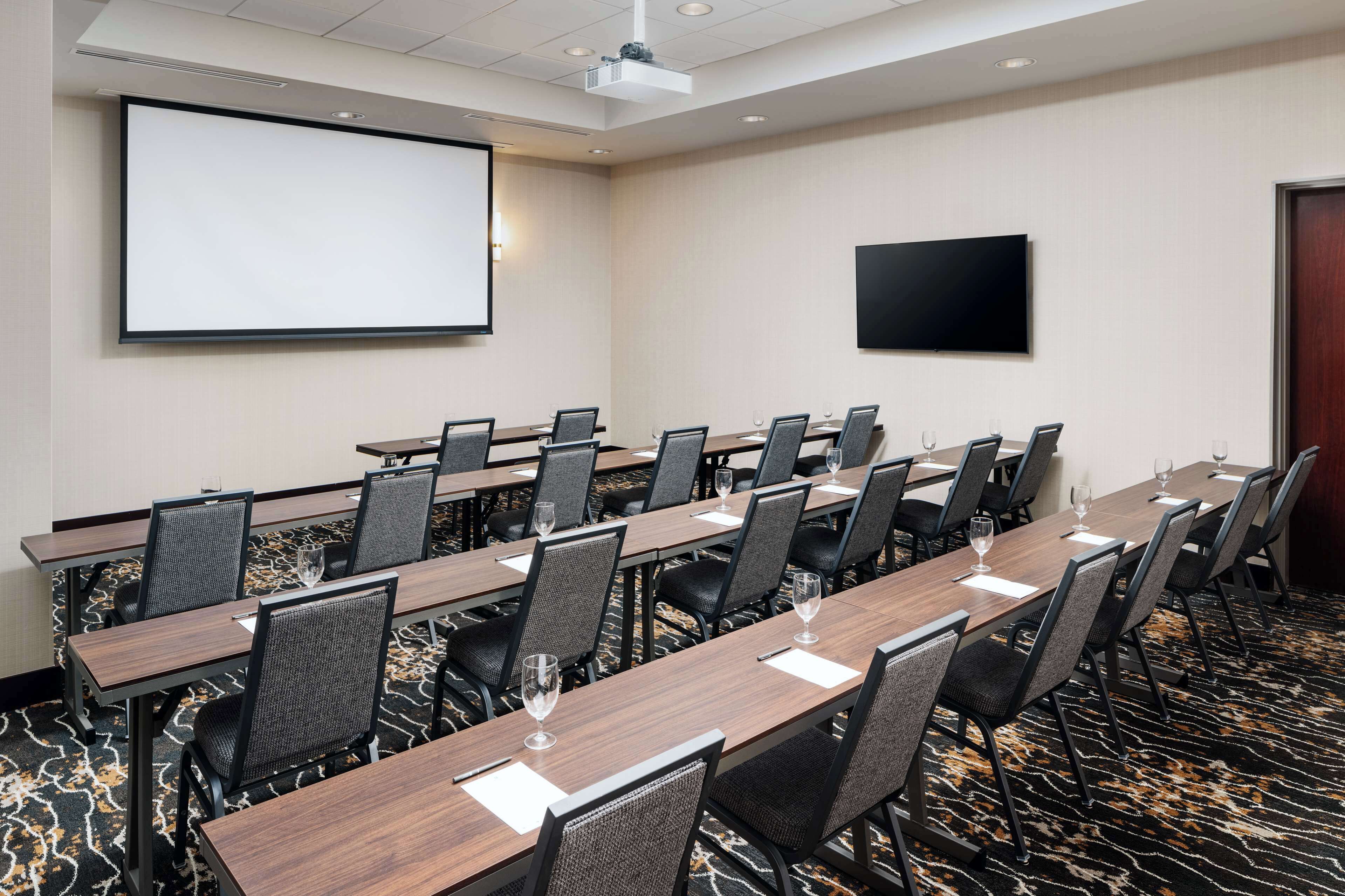 A conference room with long tables, chairs, a projector screen, and a flat-screen TV.