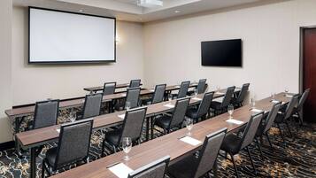 A conference room with long tables, chairs, a projector screen, and a flat-screen TV.