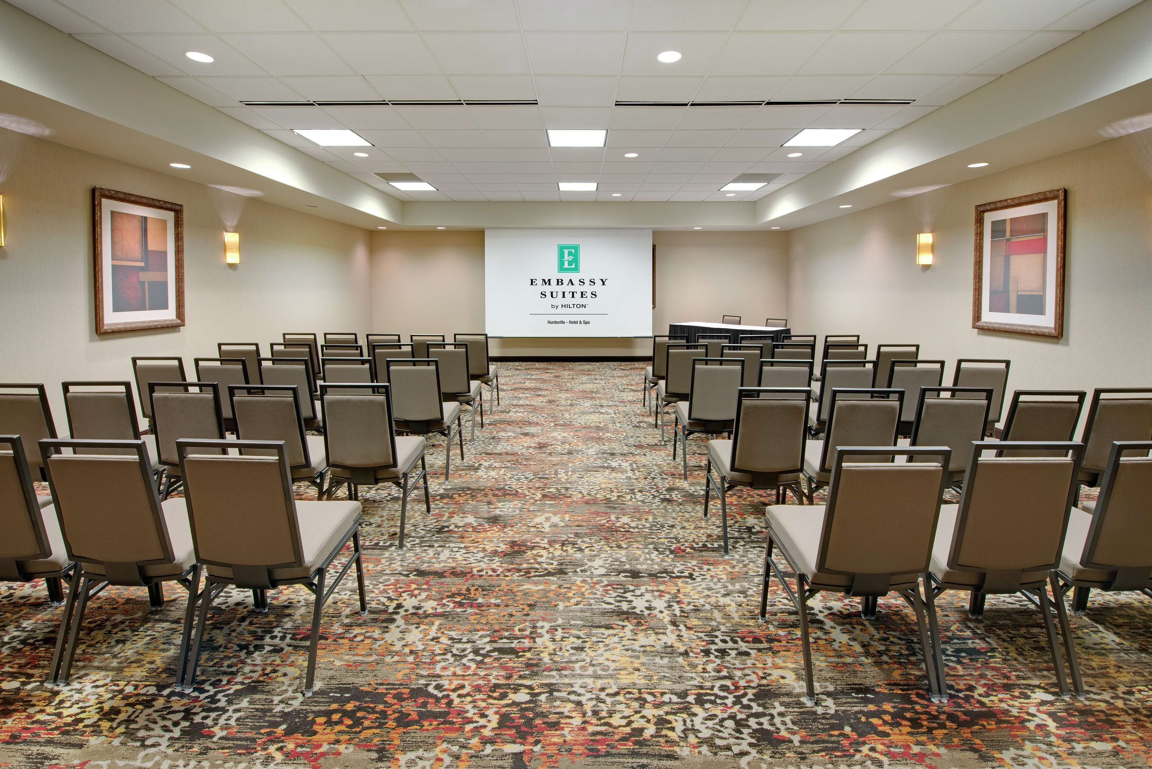 Un salón de conferencias con filas de sillas, piso alfombrado y una pantalla mostrando 'Embassy Suites by Hilton'.