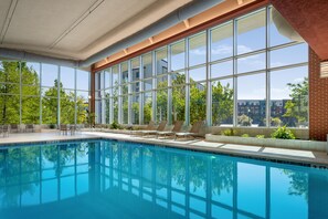 Una piscina con agua azul cristalina, rodeada de sillas de descanso y vistas a árboles y edificios a través de amplios ventanales.