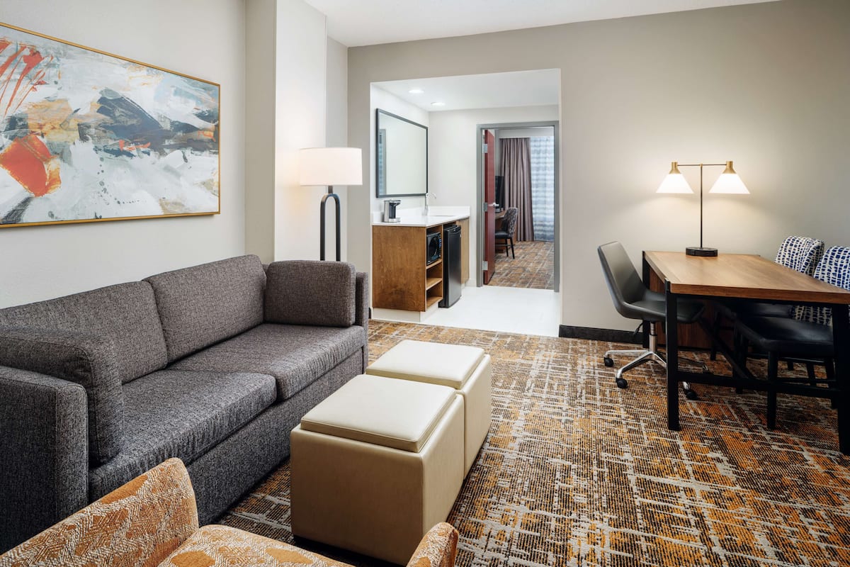 Une chambre d’hôtel moderne avec un canapé gris, des poufs, une table à manger avec des chaises, une lampe et un grand tableau abstrait accroché au mur.