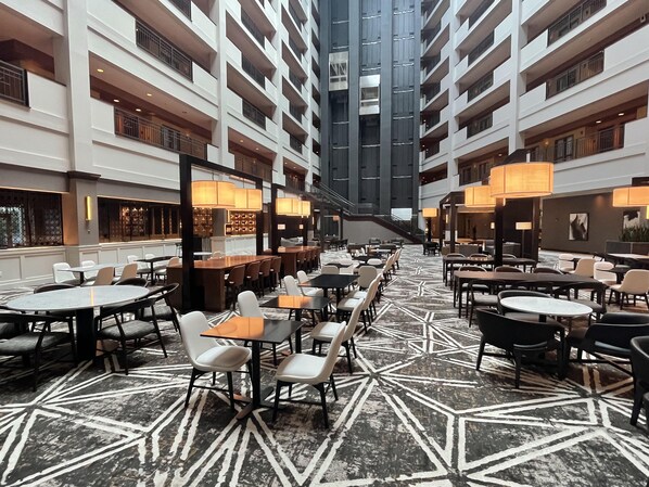 Un vestíbulo de hotel con varias zonas de comedor, una escalera central y un techo alto.
