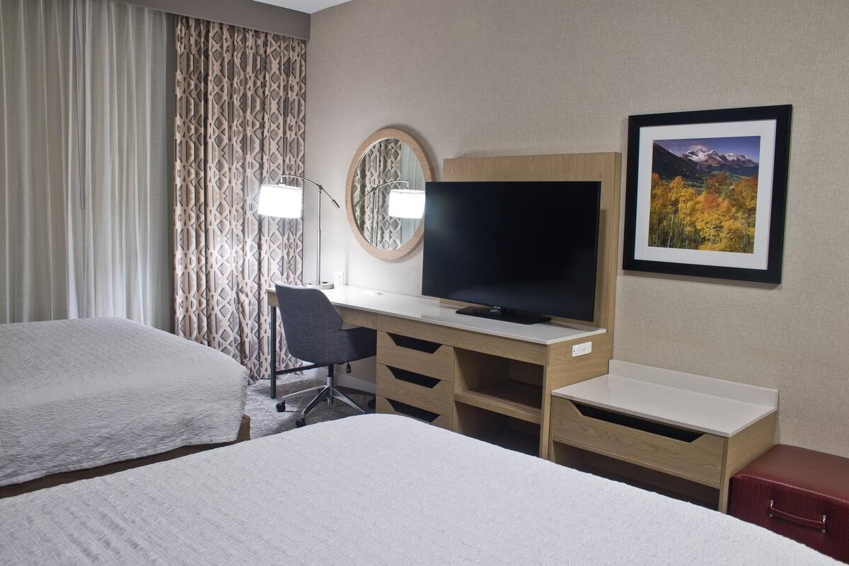 A hotel room with two beds, a desk with a TV, a chair, a small table, and a red suitcase.