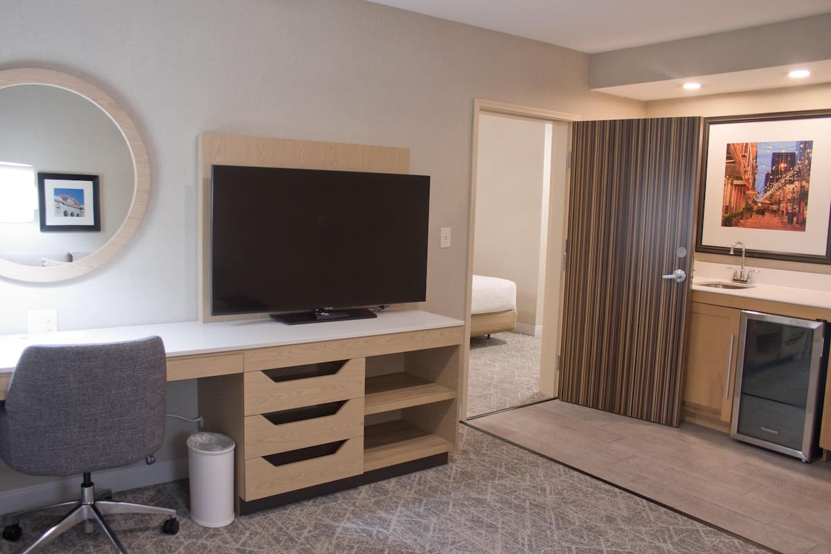 A hotel room with a wooden desk, a television, a small refrigerator, a bathroom with a shower, and a framed picture on the wall.