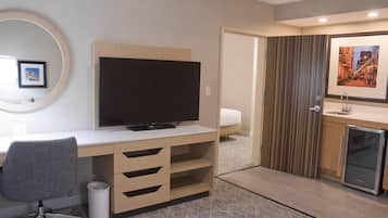Habitación de hotel con escritorio de madera, televisor, refrigerador pequeño, baño con ducha y un cuadro en la pared.