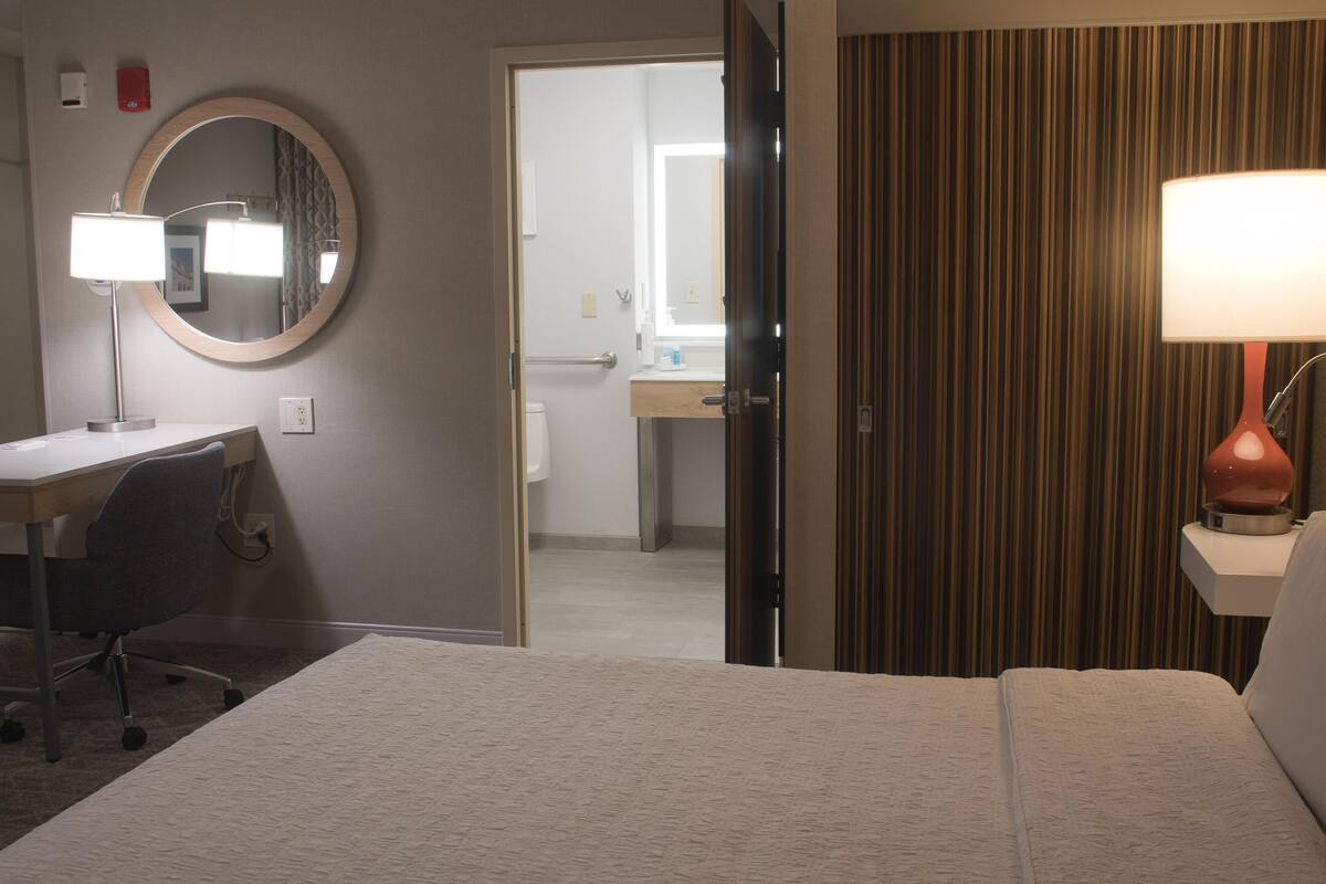 A hotel room with a bed, a desk with a chair, a round mirror, and a bedside lamp.