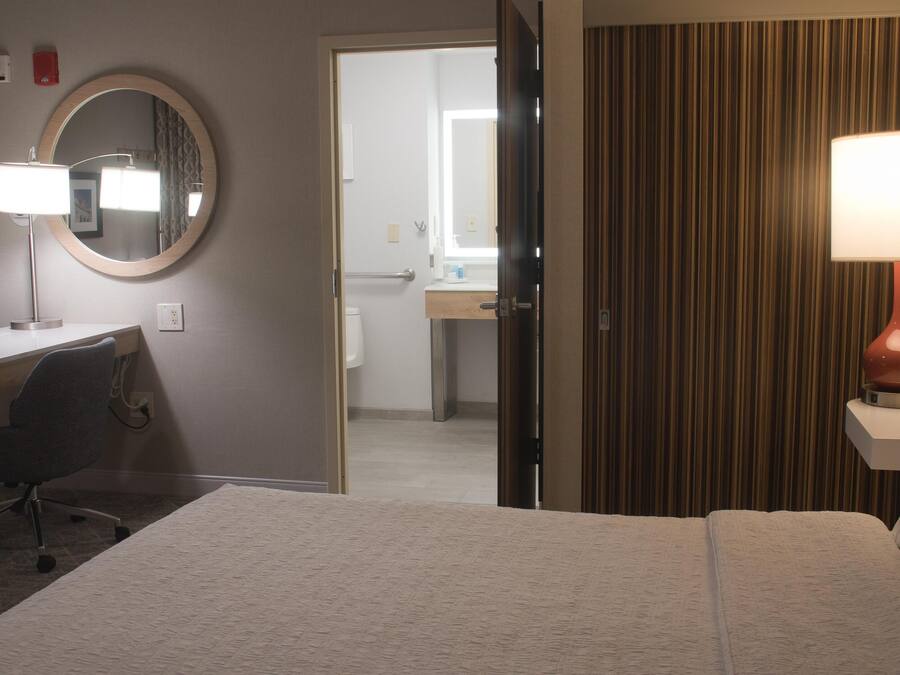 Quarto de hotel com uma cama, uma escrivaninha com cadeira, um espelho redondo e um abajur.