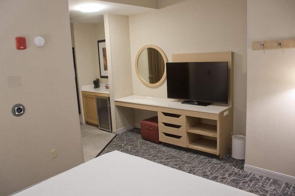 A hotel room with a desk, television, mirror, and a small red suitcase.