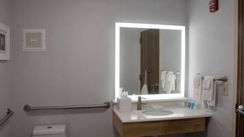 A modern bathroom with a white sink, a mirror with a light, a towel rack, and a handrail.