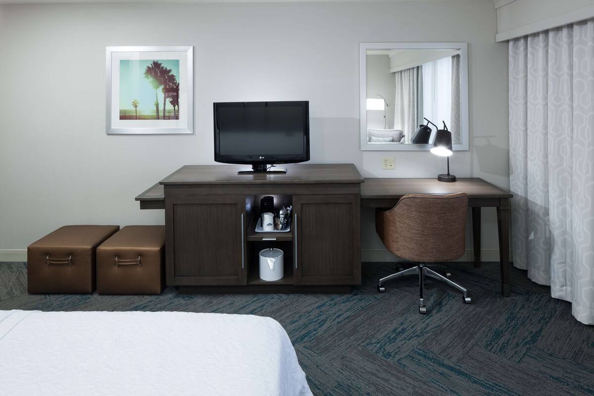 A hotel room with a bed, a desk with a computer monitor, a chair, a mirror, a framed picture on the wall, and two brown suitcases.
