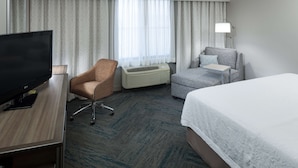 A hotel room with a bed, a desk with a TV, a chair, an air conditioner, and a lamp. - Hampton Inn Los Angeles-Orange County-Cypress (Cypress)