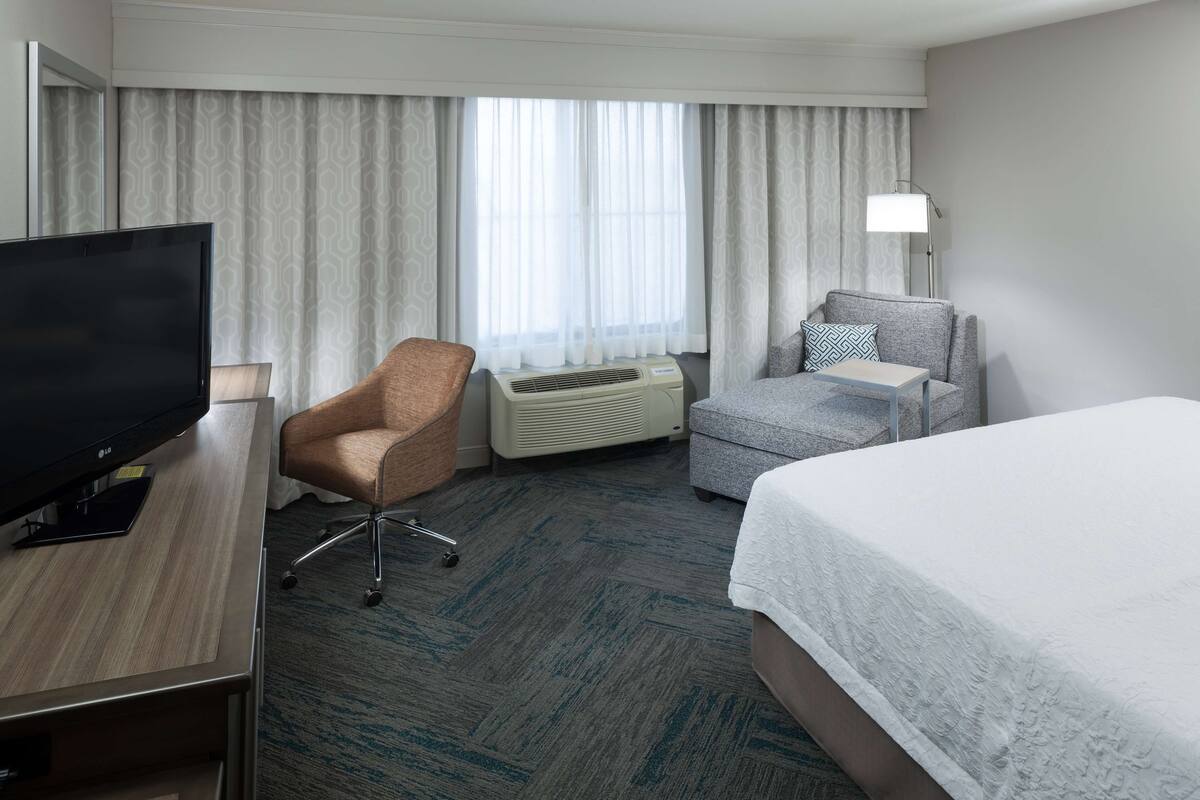 A hotel room with a bed, a desk with a TV, a chair, an air conditioner, and a lamp.