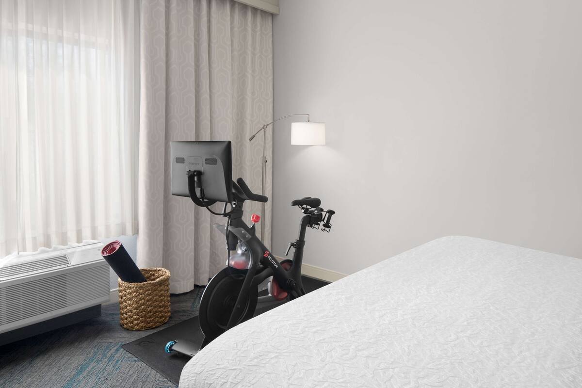 A hotel room with a bed, a standing fan, a wall-mounted heater, a bicycle, and a lamp.