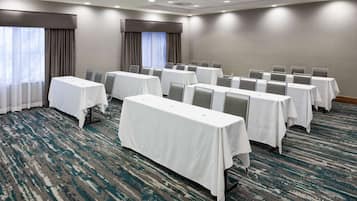 A conference room with rows of tables and chairs, carpeted floor, and large windows with curtains.