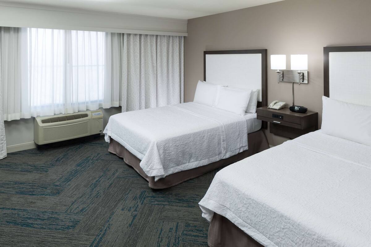 A hotel room with two beds, a bedside table with a phone and a lamp, a window with curtains, and a wall-mounted air conditioning unit.