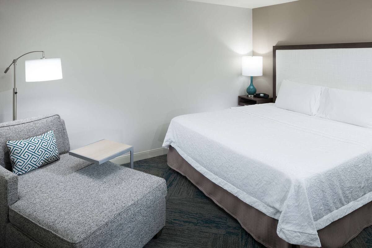 A hotel room with a bed, a chair, a nightstand, and a lamp.