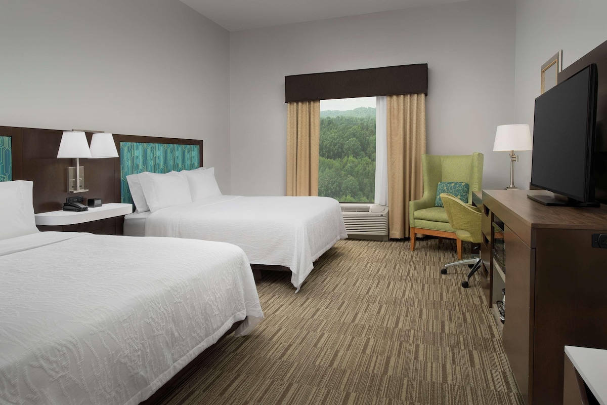 A hotel room with two beds, a television, a desk with a lamp, and a view of a forest through the window.