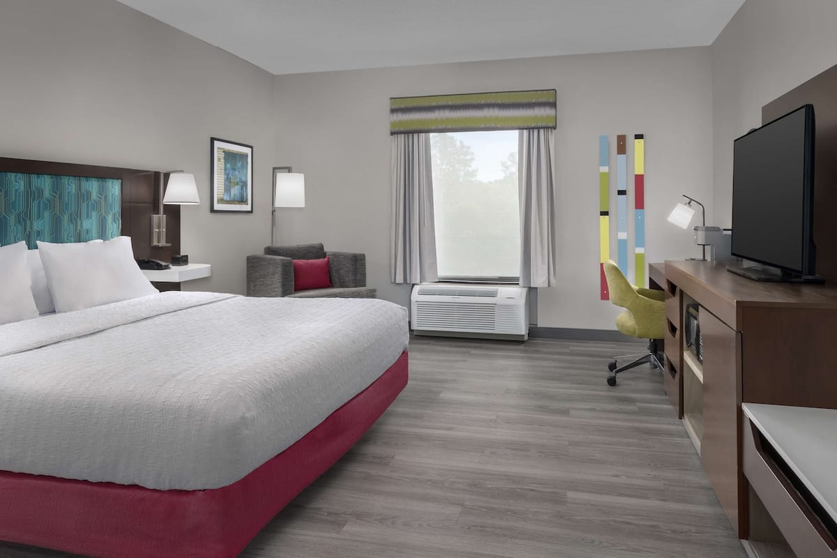 A hotel room with a large bed, a desk with a flat-screen TV, a chair, a desk lamp, an air conditioning unit, and a window with curtains.