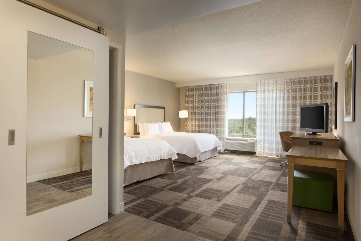 A hotel room with two beds, a desk with a television, and a large window with curtains.