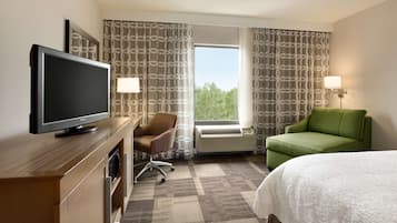 Une chambre d’hôtel équipée d’un téléviseur à écran plat, d’un fauteuil vert, d’un lit blanc et d’une fenêtre avec des rideaux à motifs.