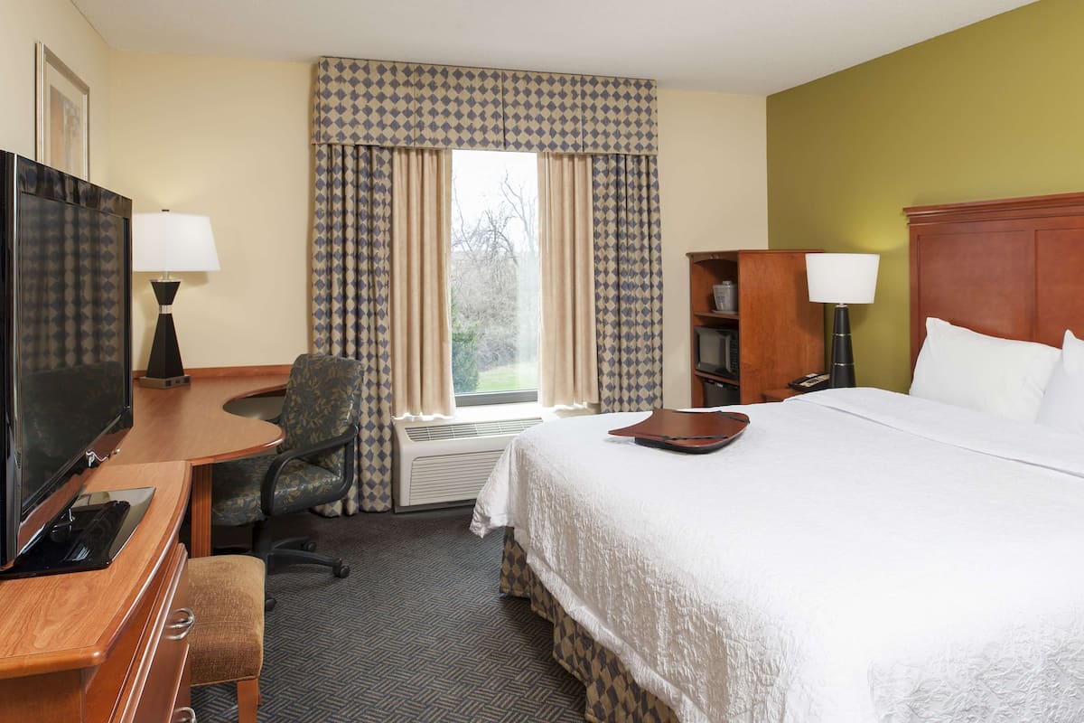 A hotel room with a bed, a desk with a TV, a chair, a bookshelf, and a window with curtains.