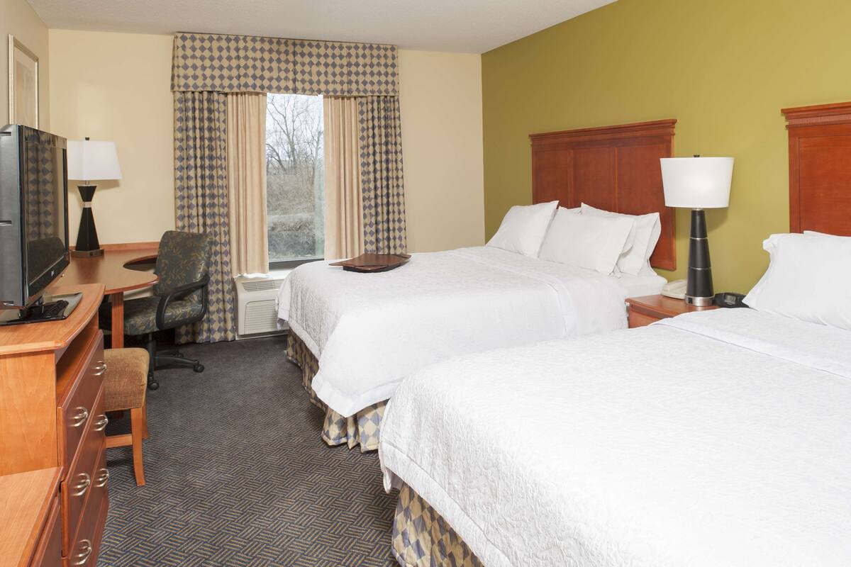 A hotel room with two beds, a desk with a television, a chair, and a window with curtains.