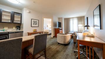 Una habitación de hotel con kitchenette, comedor, sala de estar con sofá y sillón, televisor y escritorio con lámpara.