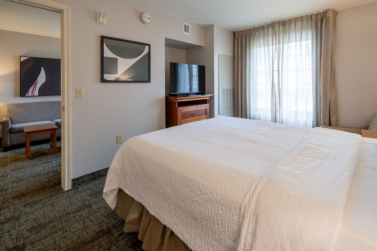 Ein Hotelzimmer mit einem großen Bett, einem Fernseher auf einem Holzständer, einem kleinen Tisch und einem Sessel.