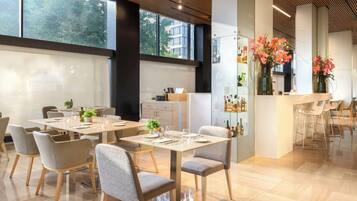 Un comedor moderno con varias mesas y sillas, una zona de bar con taburetes, y vistas de la ciudad a través de grandes ventanales.