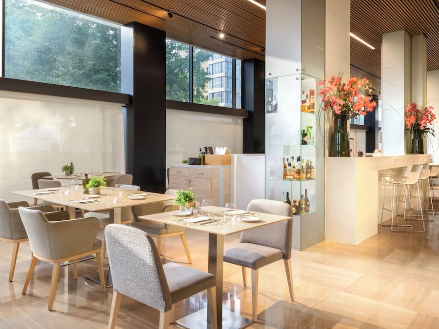 Un comedor moderno con varias mesas y sillas, una zona de bar con taburetes, y vistas de la ciudad a través de grandes ventanales.