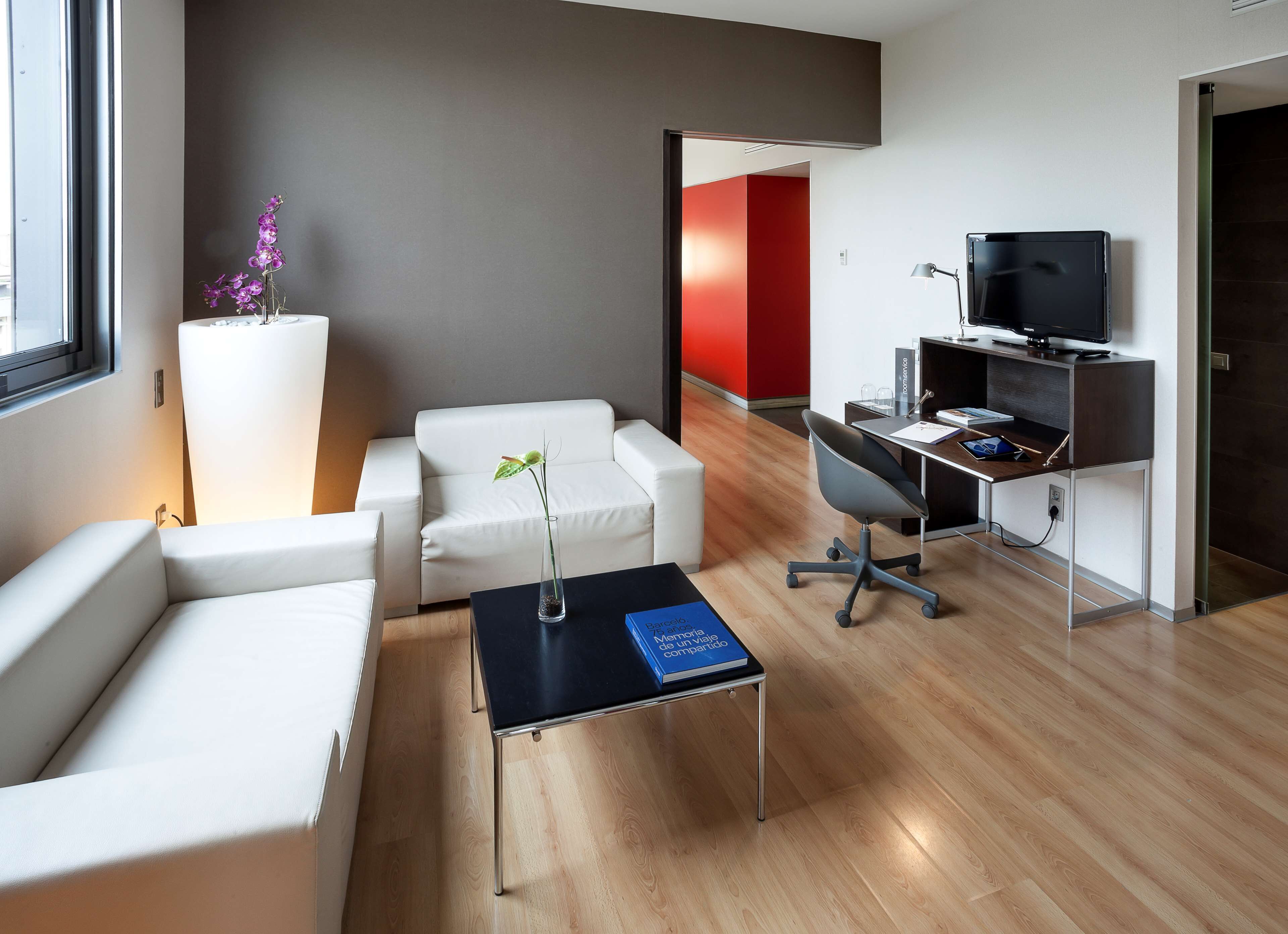A modern hotel room with a white sofa, a small coffee table, a desk with a computer and TV, and a large window.