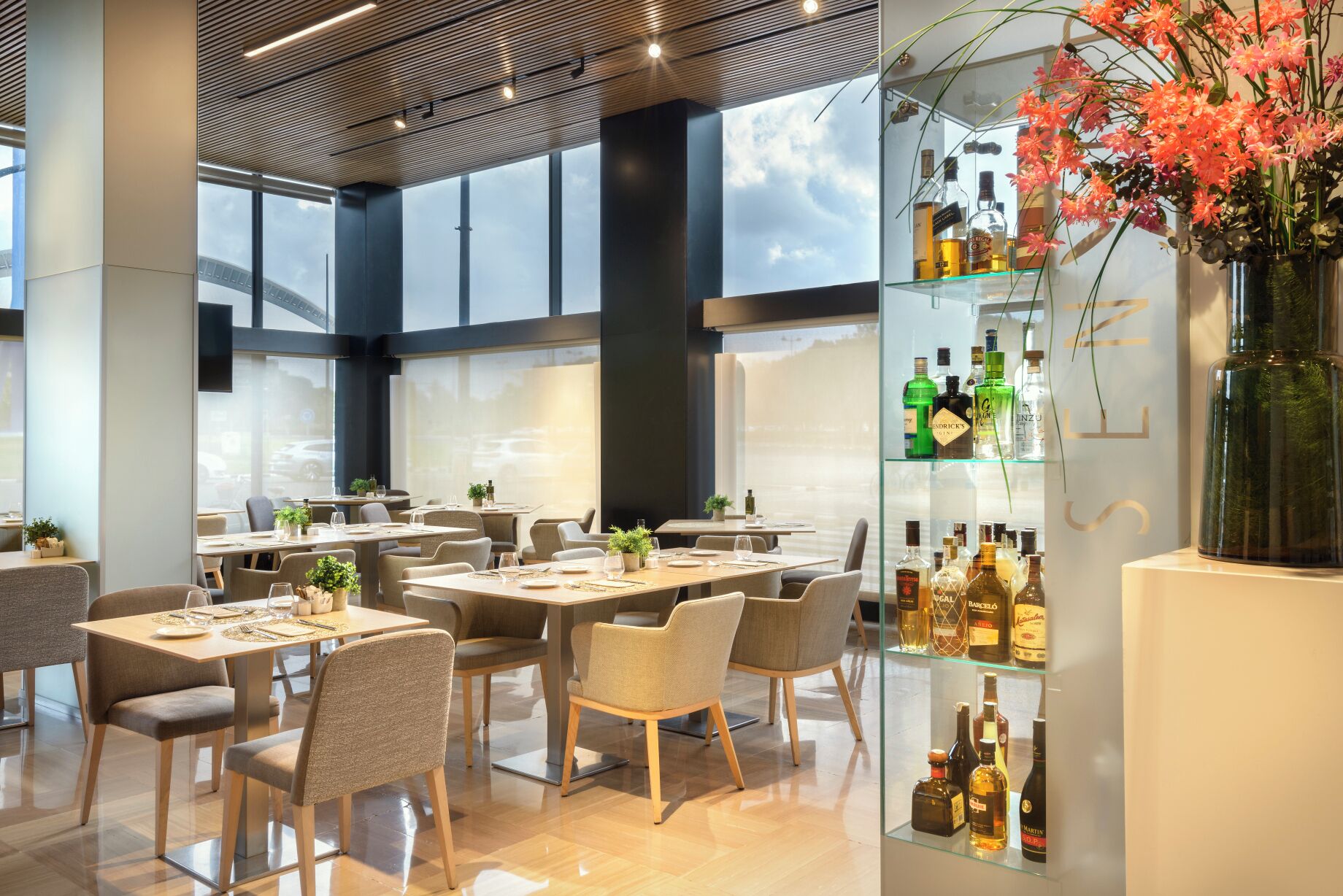 A modern restaurant interior with high ceilings, large windows, and a bar area with various liquor bottles.