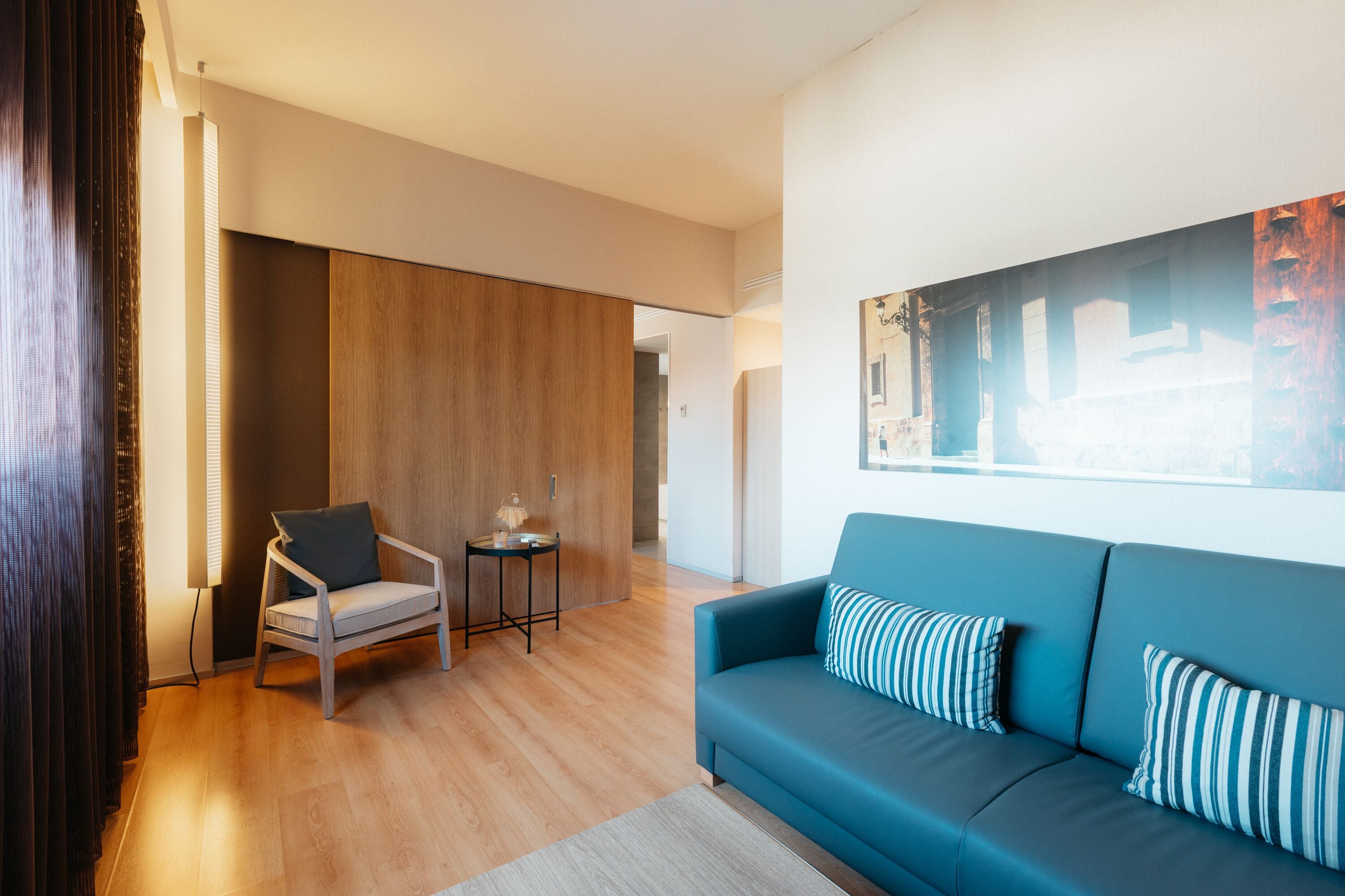 A modern living room with a blue sofa, a wooden floor, a small table with a lamp, and a large window with blinds.