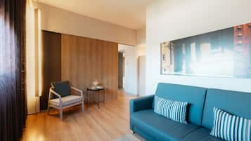 Un salón moderno con un sofá azul, suelo de madera, una mesita con lámpara y un gran ventanal con persianas.