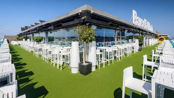 Una terraza en la azotea con mesas y sillas blancas, paneles solares y fachada de vidrio.