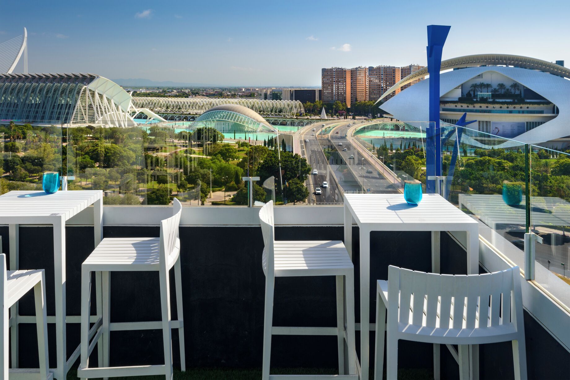 A balcony with white chairs and tables, overlooking a cityscape with modern architecture and a highway.